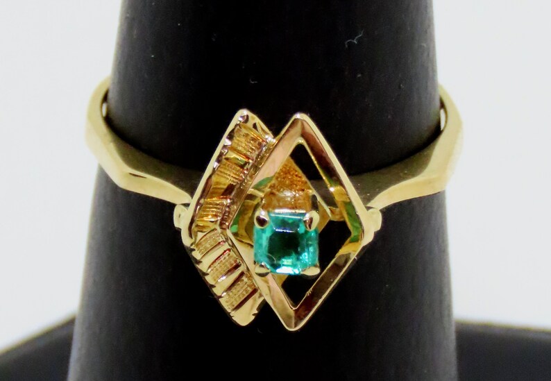 May include: A gold ring featuring a square emerald gemstone set within a diamond-shaped frame. The ring has a textured design on one side and a smooth, polished band. The emerald is a vibrant green color.