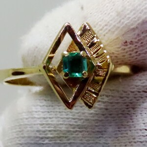 May include: A gold ring featuring a square emerald gemstone set within a geometric diamond-shaped design. The ring has a thin band and intricate detailing on the side of the diamond shape. The emerald is a vibrant green color.