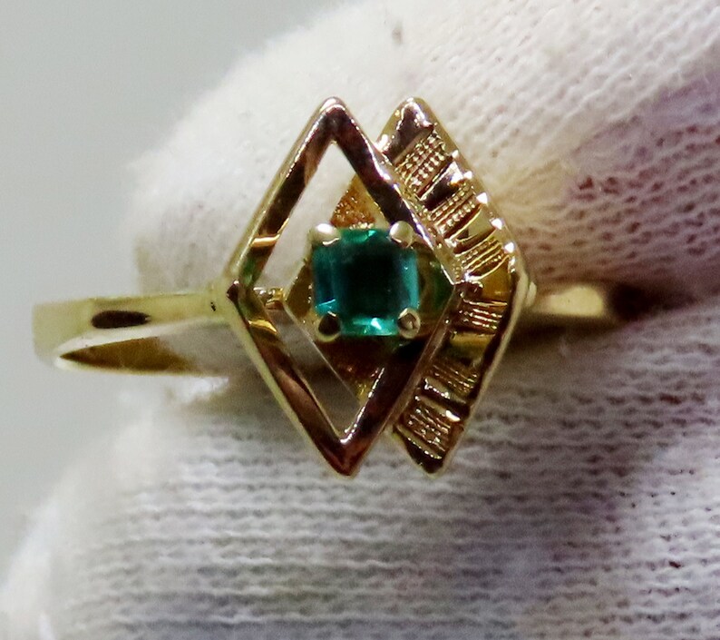 May include: A gold ring featuring a square emerald gemstone set within a geometric diamond-shaped design. The ring has a thin band and intricate detailing on the side of the diamond shape. The emerald is a vibrant green color.