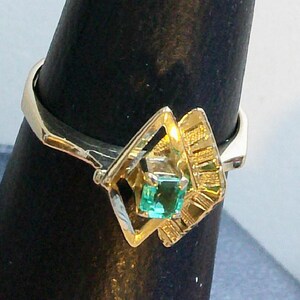 May include: A gold ring featuring a square, emerald-cut green gemstone. The ring has a unique geometric design with a diamond-shaped setting and textured gold accents. The ring is displayed on a black stand.