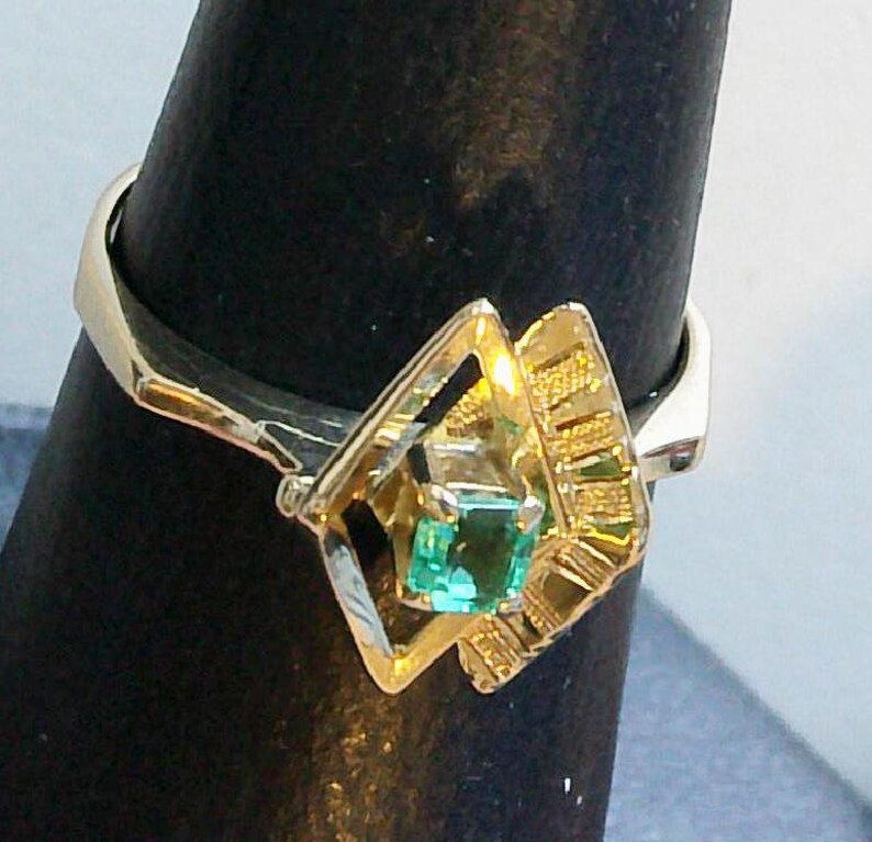 May include: A gold ring featuring a square, emerald-cut green gemstone. The ring has a unique geometric design with a diamond-shaped setting and textured gold accents. The ring is displayed on a black stand.