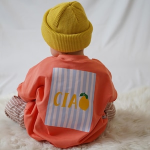 Könnte beinhalten: Ein Kind trägt eine senfgelbe Mütze und ein korallenfarbenes Langarmshirt. Das Shirt hat ein rechteckiges Design mit blau-weißen Streifen, das Wort "CIAO" in Gelb und eine gelbe Zitronenillustration.