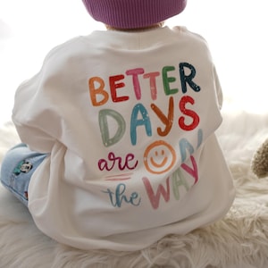 Könnte beinhalten: Ein weißes Sweatshirt mit dem Text "BETTER DAYS are the WAY" in bunten Buchstaben und einem Smiley-Gesicht. Das Sweatshirt wird von einem Kind getragen. Ein Teddybär ist im Hintergrund.