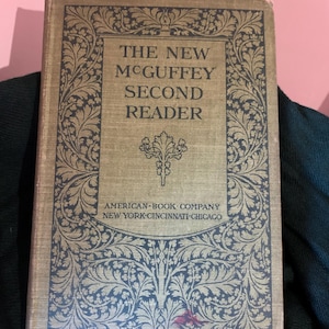 Puede incluir: La portada de un libro antiguo titulado "The New McGuffey Second Reader" publicado por American Book Company en Nueva York, Cincinnati y Chicago. La portada es de color marrón oscuro con un borde decorativo de diseños florales negros.