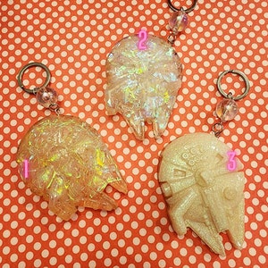 Può includere: Tre portachiavi in resina bianca iridescente a forma di nave spaziale Millennium Falcon di Star Wars. Ogni portachiavi ha un anello portachiavi argentato e una perla di vetro trasparente.