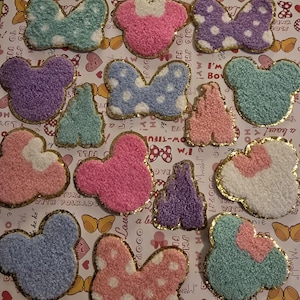 May include: A collection of 14 colorful felt patches with various shapes and designs, including Mickey Mouse ears, bows, and hearts. The patches are outlined in gold glitter and have a fuzzy texture.