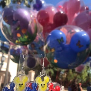 Pu&ograve; includere: Un paio di orecchini pendenti con tre palloncini a forma di testa di Topolino in rosso, giallo e blu. I palloncini sono all'interno di plastica trasparente e hanno un gancio argentato.
