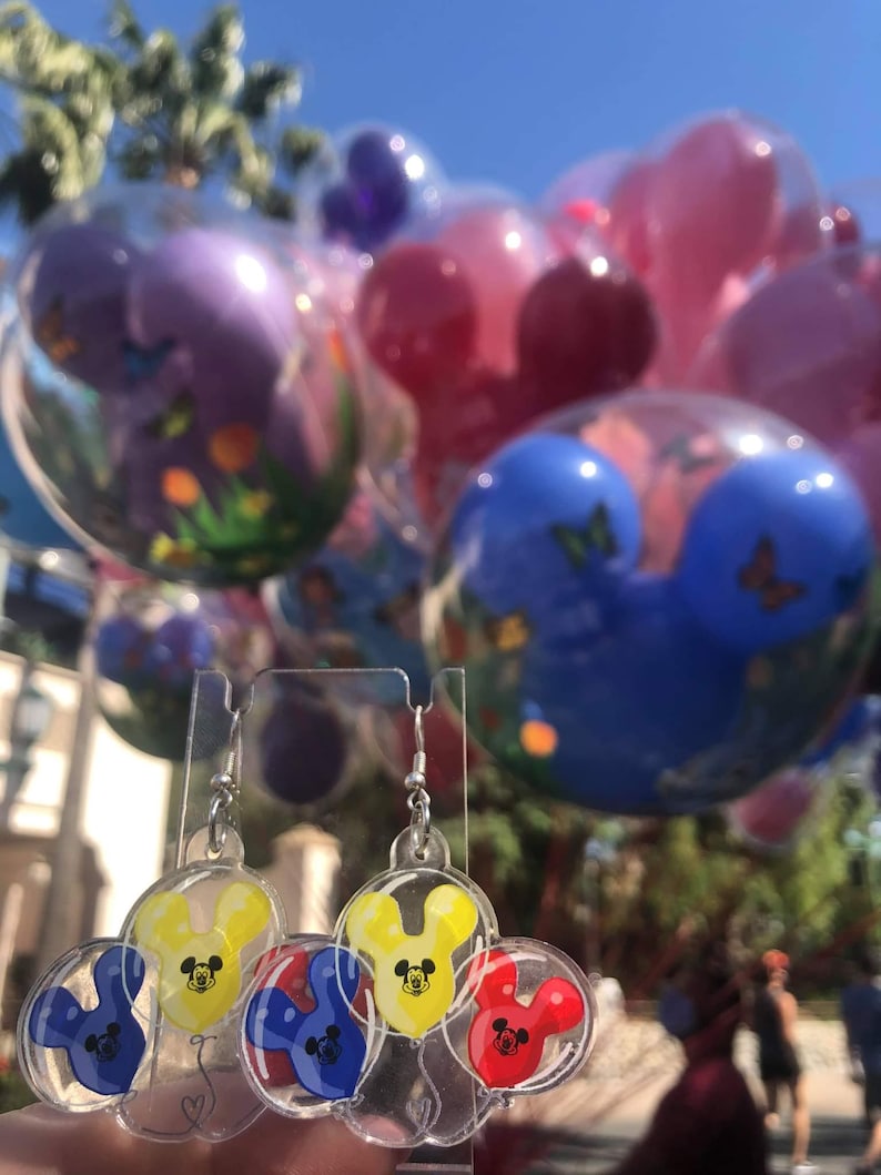 Pu&ograve; includere: Un paio di orecchini pendenti con tre palloncini a forma di testa di Topolino in rosso, giallo e blu. I palloncini sono all'interno di plastica trasparente e hanno un gancio argentato.