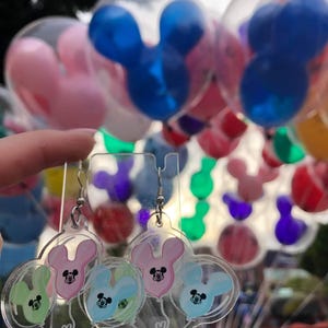 Pu&ograve; includere: Orecchini in acrilico trasparente con un design a forma di testa di Topolino. Gli orecchini presentano un gruppo di tre palloncini a forma di testa di Topolino in rosa, blu e verde.