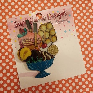 Puede incluir: Un amuleto de helado de sundae colorido con una rebanada de pastel rosa, un cono de waffle, una cereza y un tazón de helado azul. El amuleto está en una tarjeta blanca con el texto "Superfluous Designs" y "2 Extra".
