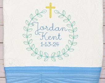 Könnte beinhalten: Eine weiße Babydecke mit blauem Rand. Die Decke hat ein gesticktes Design mit einem Kreuz, einem Kranz aus Blättern und dem Text "Jordan Kent 1-13-24".