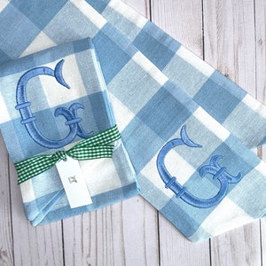 May include: Two blue and white gingham fabric napkins with a blue embroidered letter G on each. The napkins are tied together with a green ribbon and a white tag.