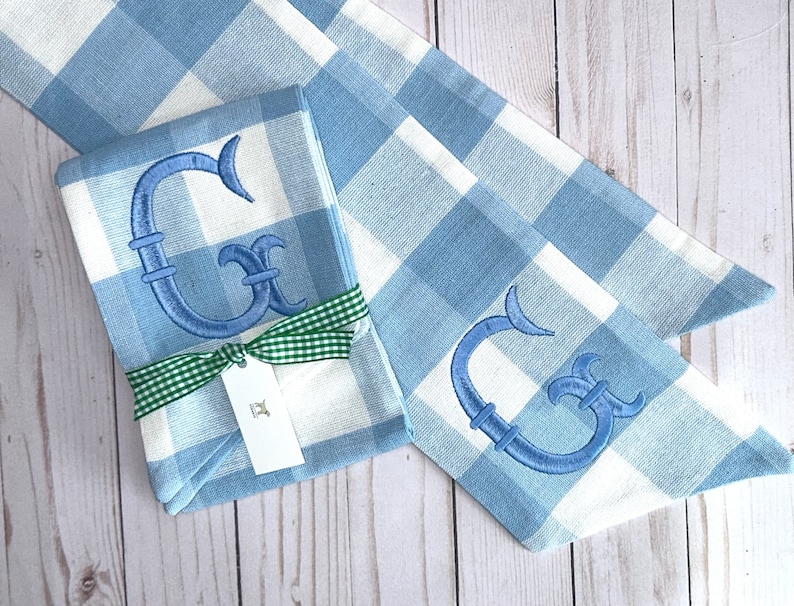 May include: Two blue and white gingham fabric napkins with a blue embroidered letter G on each. The napkins are tied together with a green ribbon and a white tag.