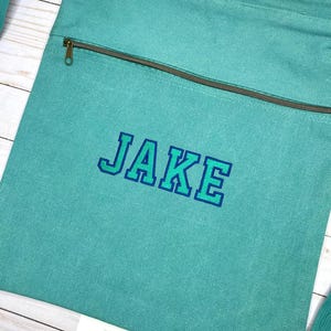 以下が含まれることがあります： ターコイズブルーのキャンバス製巾着袋。ジッパー付きポケットと、青と緑の文字で「JAKE」と刺繍されています。