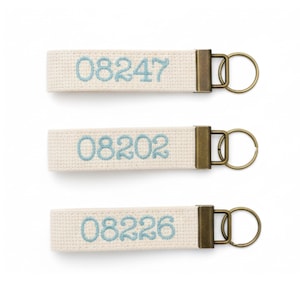 May include: Three cream-coloured keychains with embroidered numbers in light blue. Each keychain has a rectangular fabric strap with a metal clasp and a keyring. The numbers embroidered are 08247, 08202, and 08226.