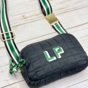 May include: Black quilted crossbody bag with a green and white striped strap. The bag features the embroidered letters "LP" in green and white. The strap has a gold-coloured buckle and a black zip.