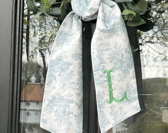 May include: A white and light blue patterned scarf with a green embroidered letter "L". The scarf is draped over a wreath with green leaves. The pattern features a pastoral scene.