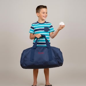 May include: A navy blue duffel bag with red lettering is held by a person wearing a blue and white striped t-shirt. A baseball is held in the person's other hand. The bag has two handles and a shoulder strap.