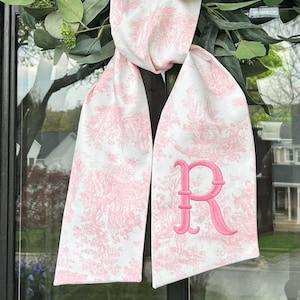 Puede incluir: Bufanda blanca con un estampado toile de Jouy rosa y una gran letra "R" rosa bordada. La bufanda está drapeada sobre una corona verde. El estampado toile de Jouy presenta una escena pastoral.