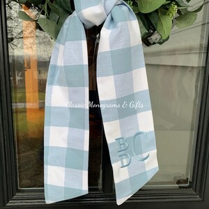 May include: A blue and white gingham ribbon with the monogram "BC" embroidered in white. The ribbon is tied in a bow and is hanging on a black door.