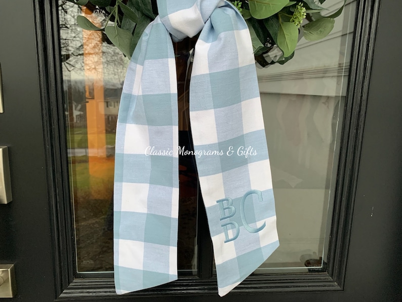 May include: A blue and white gingham ribbon with the monogram "BC" embroidered in white. The ribbon is tied in a bow and is hanging on a black door.