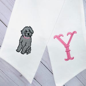 Op de afbeelding: Twee witte stoffen items met geborduurde ontwerpen. Eén toont een grijze hond met een roze halsband, de andere een roze letter "Y". De items zijn waarschijnlijk decoratief of gepersonaliseerd.