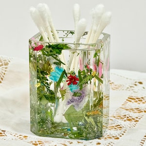 May include: A clear, hexagonal resin holder containing embedded dried green foliage, small blue and red flowers, and gold leaf flakes. Several white cotton swabs are placed upright inside the container, which has a smooth, transparent finish.