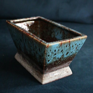 May include: A rectangular ceramic planter with a flared rim, featuring a reactive glaze that transitions from a light blue and brown mottled pattern at the top to a dark brown middle section and a textured, light beige base with a pedestal foot.