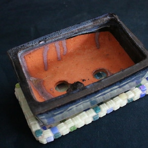 Puede incluir: Una maceta rectangular de cerámica para bonsái con exterior esmaltado en azul oscuro e interior naranja sin esmaltar con dos orificios de drenaje. La maceta descansa sobre una base rectangular decorada con una cuadrícula de pequeñas piezas de mosaico blancas, azules y verdes.