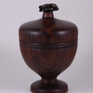 May include: A dark brown wooden lidded vessel with a smooth, polished finish and a visible wood grain pattern. The container features a rounded body, a pedestal base, and a lid topped with a small, textured, natural-looking wooden finial.