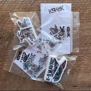 May include: Clear plastic bags containing stickers with a dog graphic. The stickers feature a dog in a tactical vest with the text "K-9 MAVRIC" and "K9&K". The stickers are on a wooden surface.