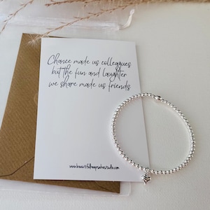 May include: A silver beaded stretch bracelet with a small heart charm, displayed next to a white card featuring the black printed text: Chance made us colleagues but the fun and laughter we share made us friends, and the website www.beautifulkeepsakestudio.com.
