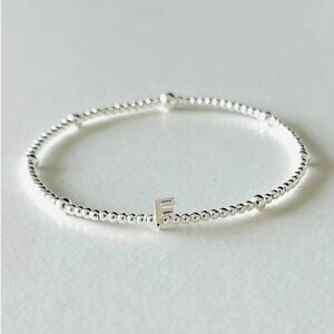 May include: A silver-toned stretch bracelet composed of small, uniform metallic beads. The bracelet features a single, prominent silver-coloured letter F charm positioned centrally among the beads, creating a simple and clean design.