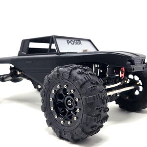 May include: Black remote control truck with large, textured tires. The truck has a cab with the word "POSER" on the windshield. The vehicle is designed for off-road use and features a detailed chassis.