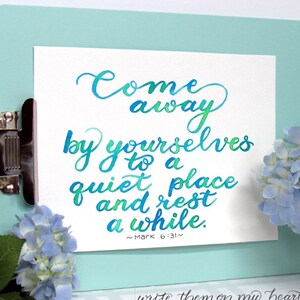 May include: A clipboard with a white paper featuring the quote "Come away by yourselves to a quiet place and rest a while." The text is in blue and green watercolor. A bouquet of blue hydrangeas sits to the right.