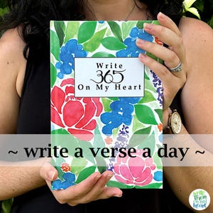 May include: A journal with a floral design featuring red, blue, and pink flowers, and green leaves. The cover has the text "Write 365 On My Heart" and "~ write a verse a day ~". The journal is held by a person.
