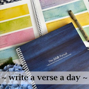May include: An open spiral-bound journal with colorful pages, a blue cover, and a black pen. The journal's pages are filled with lines and text, with the words "Write them on my heart" and "The Write Pursuit" visible. The phrase "write a verse a day" is also present.