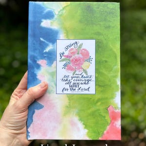 May include: Lined journal with a watercolor design in shades of blue, green, and pink. The cover features a floral illustration and the text "Be Strong and let your heart take courage all you who wait for the Lord."