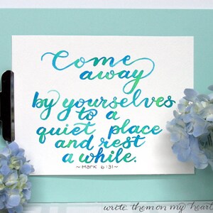 May include: A white paper with teal and green calligraphy script, clipped to a light blue clipboard. The text reads "Come away by yourselves to a quiet place and rest a while." Blue hydrangeas are in the background.