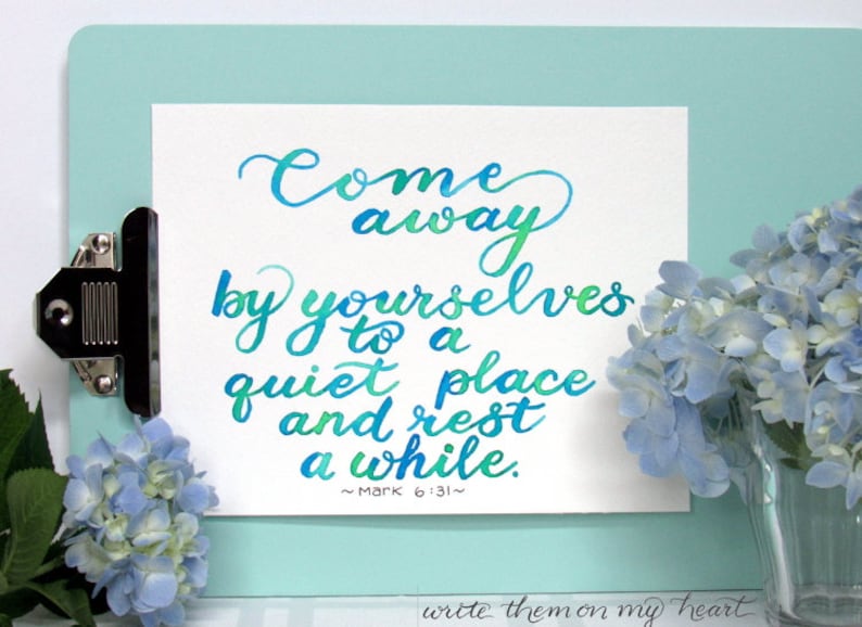 May include: A white paper with teal and green calligraphy script, clipped to a light blue clipboard. The text reads "Come away by yourselves to a quiet place and rest a while." Blue hydrangeas are in the background.