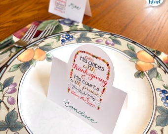 May include: A white place card with the name "Candace" in green script sits on a decorative plate. The card features the text "Enter His gates with thanksgiving & His courts with praise!" A second place card with the name "Mike" is in the background.