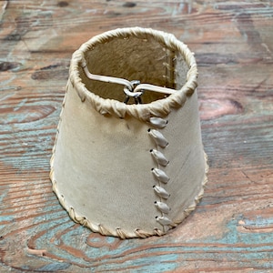 May include: A white leather lampshade with a stitched seam down the side. The shade has a metal ring at the top for hanging.