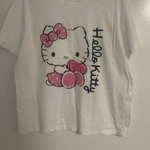 May include: White t-shirt featuring a Hello Kitty graphic. The graphic includes a pink bow with white polka dots, a pink dress, and the words "Hello Kitty" in black script. The shirt has a crew neck and short sleeves.