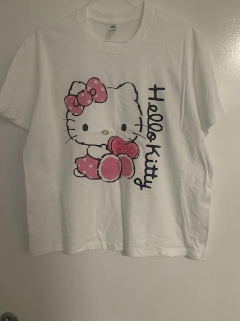 May include: White t-shirt featuring a Hello Kitty graphic. The graphic includes a pink bow with white polka dots, a pink dress, and the words "Hello Kitty" in black script. The shirt has a crew neck and short sleeves.
