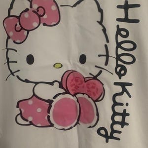 May include: White fabric featuring a Hello Kitty graphic. The design includes a pink bow with white polka dots, a pink dress with white polka dots, and the words "Hello Kitty" in black script.
