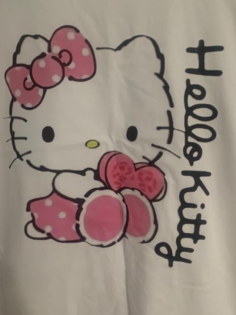 May include: White fabric featuring a Hello Kitty graphic. The design includes a pink bow with white polka dots, a pink dress with white polka dots, and the words "Hello Kitty" in black script.
