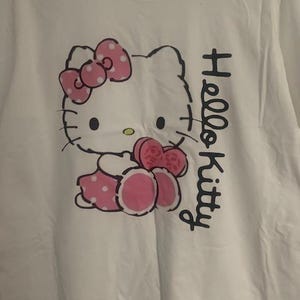 May include: White t-shirt featuring a Hello Kitty graphic. The design includes a pink bow, dress, and accents. The words "Hello Kitty" are printed vertically in black on the right side of the shirt.
