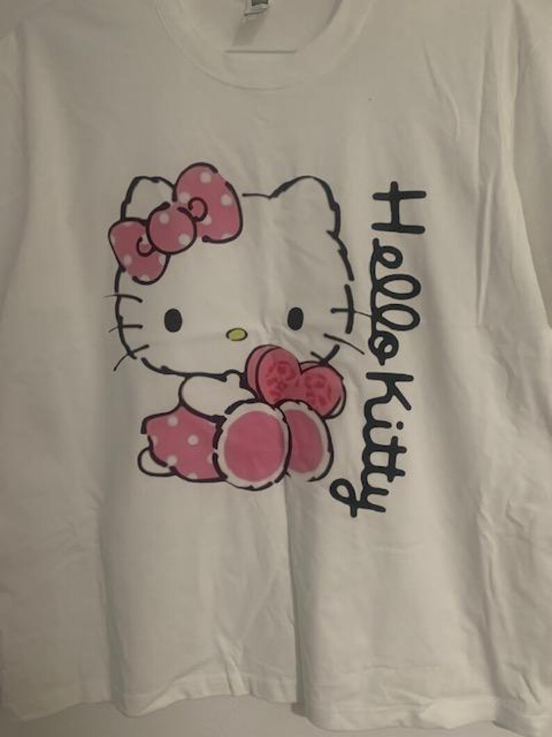 May include: White t-shirt featuring a Hello Kitty graphic. The design includes a pink bow, dress, and accents. The words "Hello Kitty" are printed vertically in black on the right side of the shirt.