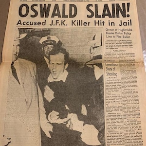 May include: A black and white newspaper headline announcing the death of Lee Harvey Oswald, the accused assassin of President John F. Kennedy. The headline reads "OSWALD SLAIN! Accused J.F.K. Killer Hit in Jail". The newspaper is dated Monday, November 23, 1963.