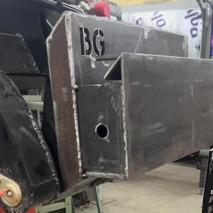 Puede incluir: Un primer plano de un accesorio de metal para una pieza de maquinaria pesada. El accesorio es gris y tiene un agujero en el centro. Las letras "BG" son visibles en el accesorio.